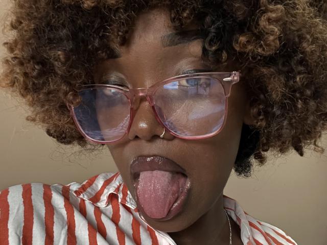 JessesXy with curly brown hair, pink glasses, nose ring, and tongue out, wearing a red and white striped shirt, showing her tongue and glossy lips.