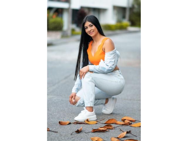 EstefaniaA with long black hair, crouching, wearing an orange top, off-shoulder blue shirt, white jeans, and white sneakers, with brown leaves on the gray pavement.