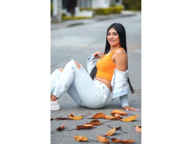 EstefaniaA with long black hair, orange crop top, white ripped jeans, and white sneakers, sitting on asphalt, jacket off, smiling, with fallen leaves around her.