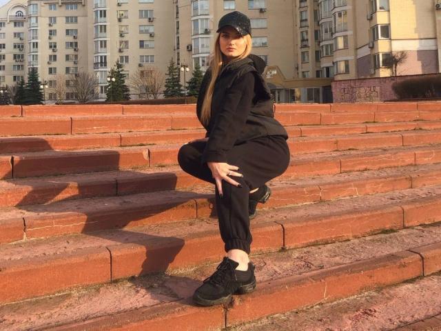 SexyCandyLove69 with long blonde hair, black cap, black hoodie, black pants, and black sneakers, squatting on red brick steps, hands on knees, looking at camera.