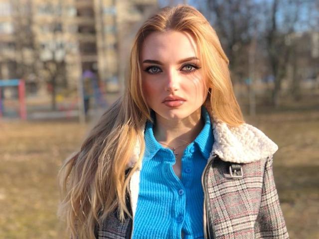 SexyCandyLove69 with long blonde hair, blue button-up shirt, and gray plaid jacket with white fur, standing outdoors, looking directly at camera, soft sunlight, slightly blurred background.