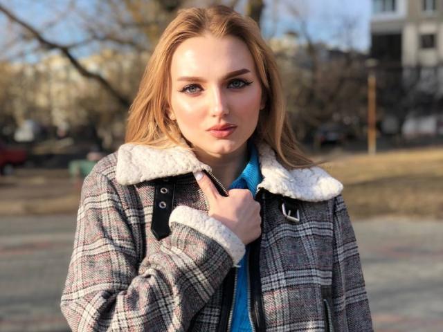 SexyCandyLove69 with blonde hair, blue eyes, and a cute face, wearing a plaid jacket with a white fur collar, holding the jacket's zipper, in a sunlit outdoor setting.