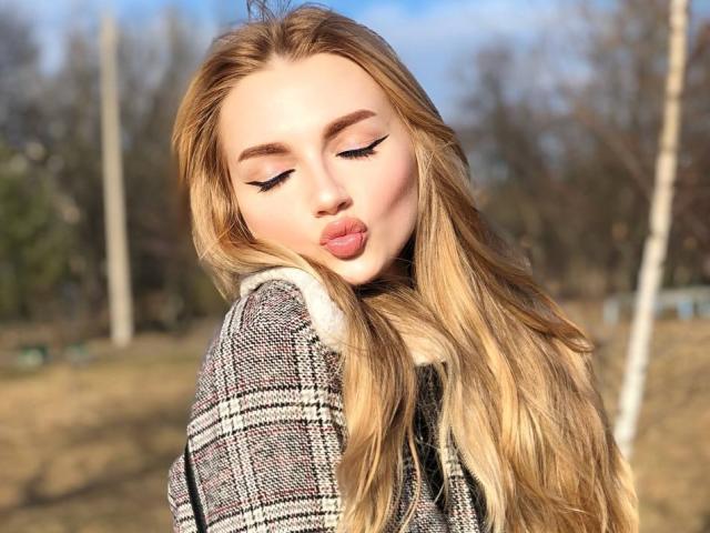 SexyCandyLove69 with long blonde hair, pouty pink lips, and winged black eyeliner, wearing a plaid coat, looking down with eyes closed, in a sunny outdoor setting.