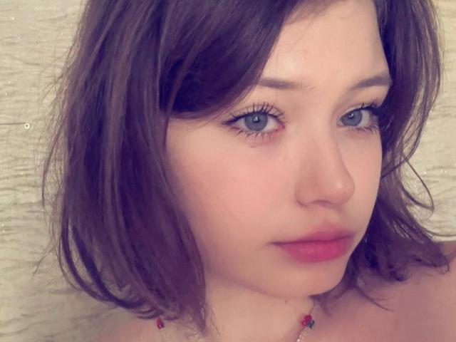 NikolKitt has a soft, pinkish blush on her cheeks, closed eyes, and a subtle, content smile. She's wearing a pink top with a bead necklace.
