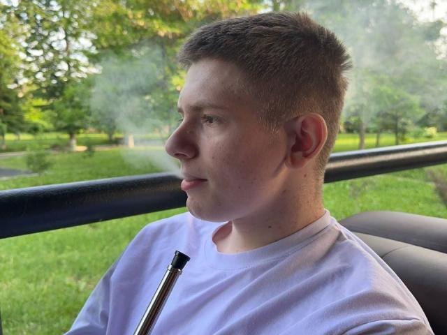 JimmieHot has a pimple on his forehead, short brown hair, black shirt, white earbuds, and sunglasses on his head, with a neutral expression, standing in a parking lot.
