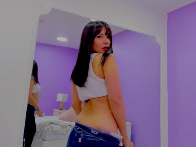 PaulinaSunny with glasses, red lipstick, and an orange off-shoulder shirt, showing her bare shoulder and smiling softly. Blue string lights in the background.