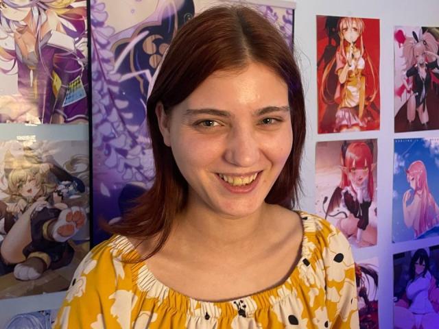 EvelynMuse met bruin haar, lachend, gekleed in een gele bloemenjurk, staand voor anime posters, met zichtbare tanden, en met een schattig, lichtelijk mollig gezicht..