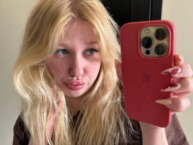 JuliKisss, loira, com lábios rosados e volumosos, olhos azuis e unhas longas, segurando um iPhone vermelho, tirando uma selfie. Seus seios estão levemente visíveis sob uma camisa marrom..