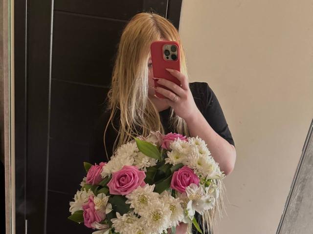 JuliKisss con i capelli biondi, una maglietta nera, che tiene rose rosa e crisantemi bianchi, sta scattando un selfie allo specchio con un telefono rosso, visibile dalla vita in su..