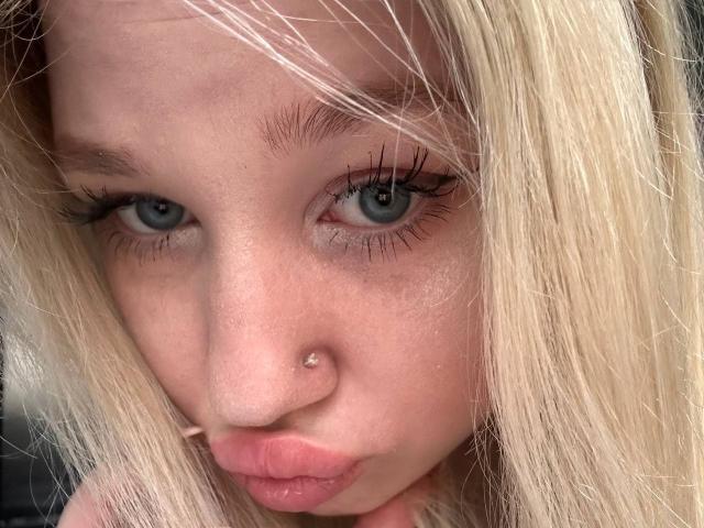 JuliKisss ha i capelli biondi, gli occhi azzurri e un piccolo piercing al naso. Sta facendo la smorfia con le sue labbra rosa. Le sue lunghe ciglia e la sua pelle chiara sono visibili..