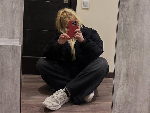 JuliKisss sitzt auf dem Boden und macht ein Selfie mit einem roten Handy, sie trägt eine schwarze Jacke, schwarze Hosen und weiße Turnschuhe, und hat langes blondes Haar..