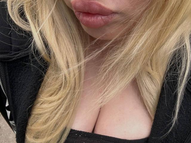 JuliKisss ha i capelli biondi, labbra piene di colore rosa e indossa una giacca nera con zip che mette in mostra il suo décolleté. Il suo seno è leggermente visibile sotto una maglia nera..