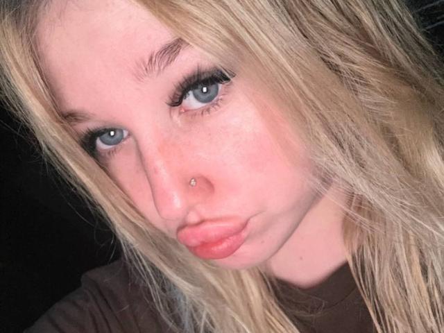 JuliKisss tiene ojos azules, un piercing en la nariz y labios rosados y gruesos, con un pequeño grano en la mejilla derecha. Su cabello rubio está despeinado y lleva una camisa marrón..