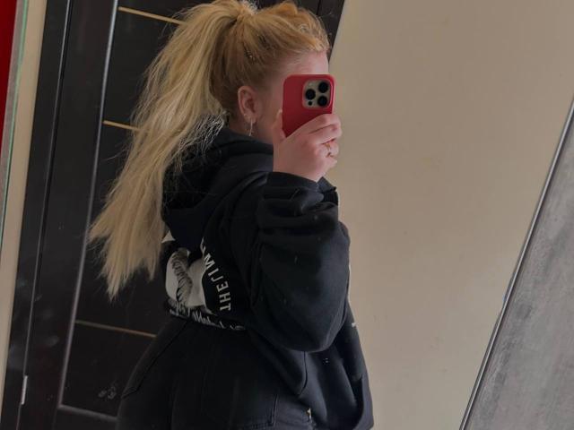 JuliKisss with blonde ponytail, black hoodie, holding red phone, covering face, taking mirror selfie, visible ass, medium build, fair skin, standing in front of dark door.