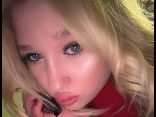 JuliKisss hat blondes Haar, blaue Augen und rosafarbene Lippen. Sie schaut mit leicht zusammengepressten Lippen in die Kamera, ihre rechte Hand berührt ihr Haar. Sie trägt ein rotes Oberteil..