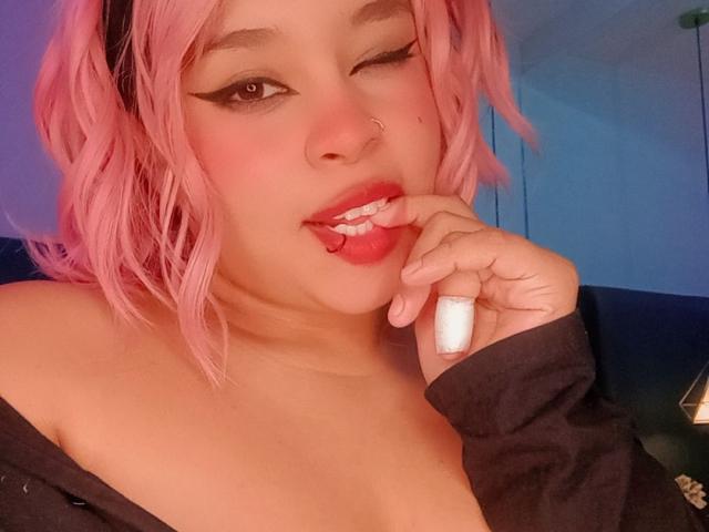 NatieRush with pink hair, winking, licking her finger, holding a white ring, wearing black off-shoulder shirt, nose ring, and winged eyeliner. Her tits are partially visible.