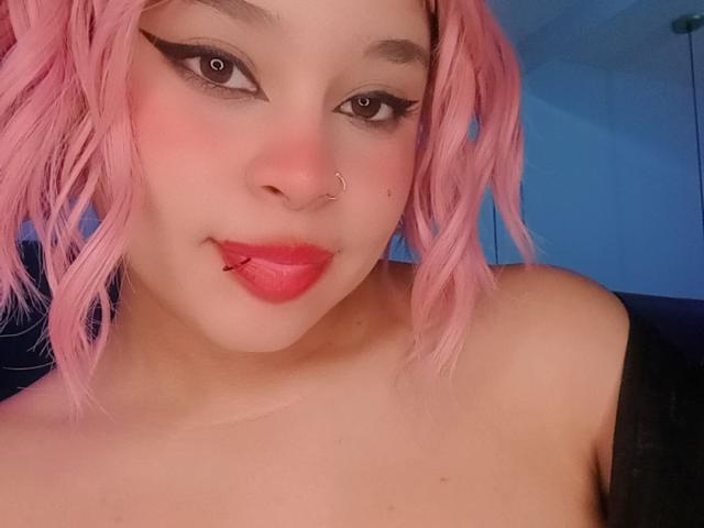 NatieRush with pink hair, nose ring, and lip ring, has black eyeliner, red lipstick, and a septum piercing, looking at the camera with a slight smile.