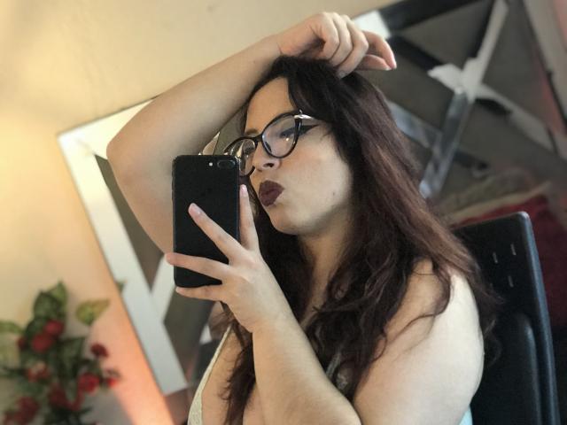Rosalinnes with glasses, pouty lips, and long brown hair, holding a phone, arm raised, showing her bare tits, in a mirror selfie. Her ass is partially visible.