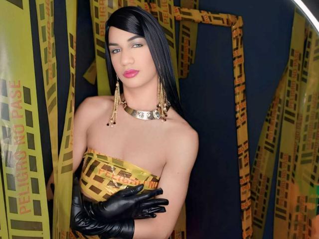 KendallSky with black hair, pink lips, gold strapless top, black gloves, gold necklace, and dangling earrings, standing against yellow and black patterned curtains. Boobs are visible.