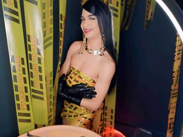 KendallSky with black gloves, gold patterned dress, and necklace, showing tits, smiling, standing in front of yellow and black striped curtains, with long black hair.