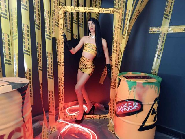 KendallSky stands in a doorway with yellow caution tape, wearing a gold, strapless, and skirted outfit, black gloves, and high heels, with long black hair and red lipstick.