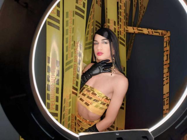 KendallSky with long black hair, wearing yellow and black checkered tube top, black gloves, and red lipstick, standing in front of patterned curtains, looking at camera.