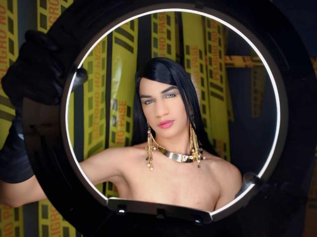 KendallSky, shirtless with black gloves, wearing gold earrings and a metallic choker, framed by a circular ring light, has blue eyes and pink lipstick, and black hair.