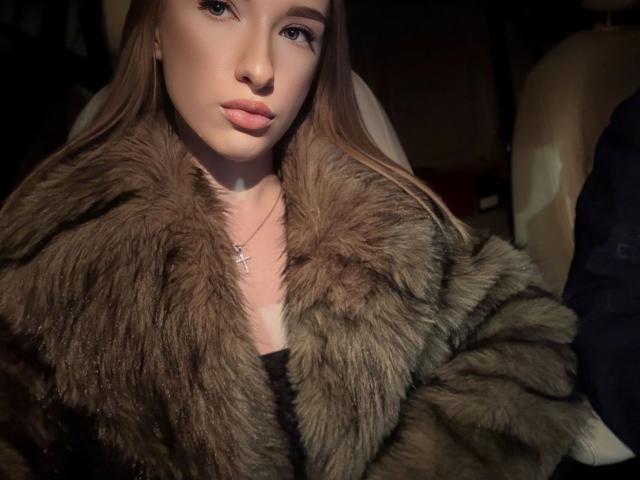 DollyVey, con lunghi capelli castani, indossa un cappotto marrone di pelliccia, una croce al collo e rossetto rosa, e guarda la fotocamera con un'espressione seria..