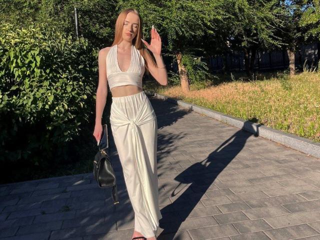 DollyVey, com cabelos loiros longos, blusa branca de alças e calças brancas de cintura alta, segurando uma bolsa preta, acenando, parque ensolarado, sombra em um caminho de tijolos, unhas dos pés rosa, magra, bonita..