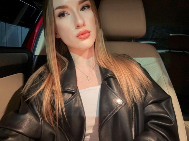 DollyVey met lang blond haar, gekleed in een zwarte leren jas, een witte top en een ketting, zittend in een auto, met een frons op haar lippen, kijkend naar de camera, zonder make-up, sexy..