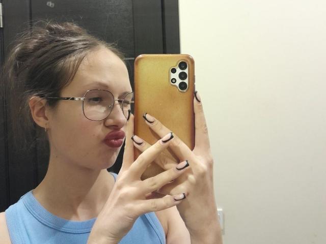 MonaCloudI con gli occhiali, labbra tumefatte e top blu, che tiene in mano un telefono dorato, mostrando unghie nere con nail art, in un selfie allo specchio..