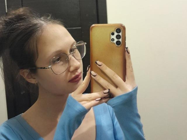 MonaCloud, che si fa un selfie con un iPhone dorato, indossando occhiali, un maglione blu e rossetto rosa, mostrando le sue unghie e un sorriso carino..