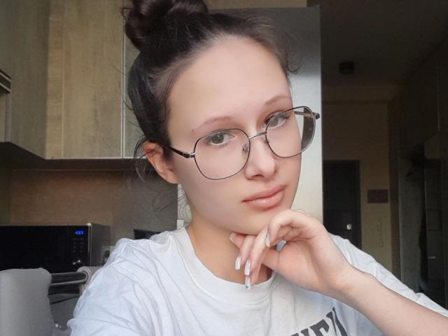 MonaCloudI, con gafas, camisa blanca y uñas largas, tiene una ligera sonrisa, la cabeza apoyada en la mano, cabello castaño recogido en un moño, piel pálida y un rostro bonito..