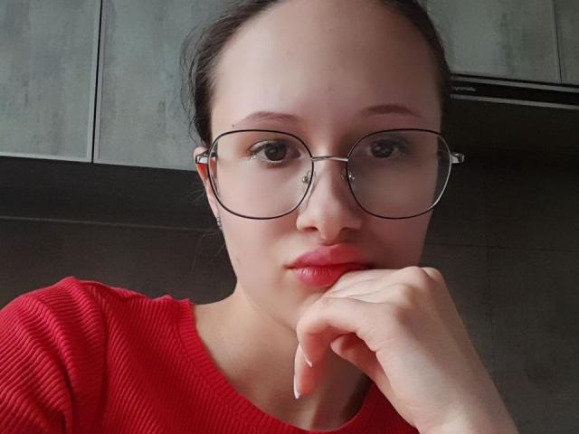 MonaCloudI, portant des lunettes, une chemise rouge à rayures et avec une main près de sa bouche, regarde directement la caméra. Elle a une peau pâle, des yeux marron et des cheveux foncés attachés en chignon..