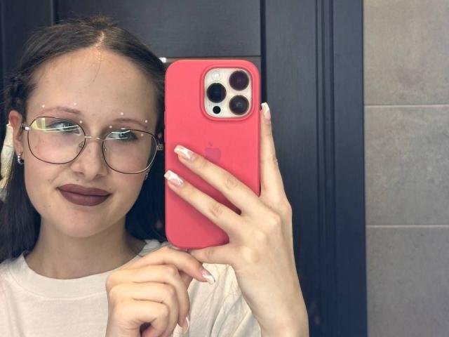 MonaCloudI con gafas, lápiz labial marrón, camisa blanca, sosteniendo un iPhone rosa, tomando una selfie, mostrando uñas cuidadas, sonriendo, en un baño, con una puerta negra y baldosas grises en el fondo..