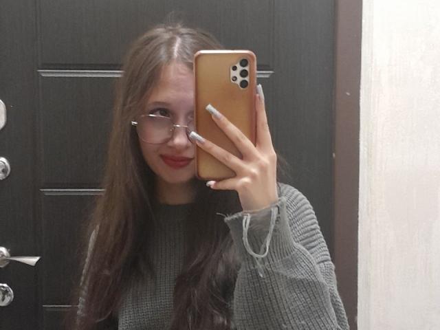 MonaCloudI, con cabello castaño largo, gafas, lápiz labial rojo y un suéter gris, sosteniendo un iPhone dorado, tomando una selfie en el espejo, mostrando sus uñas y sonriendo. No se ven los senos..
