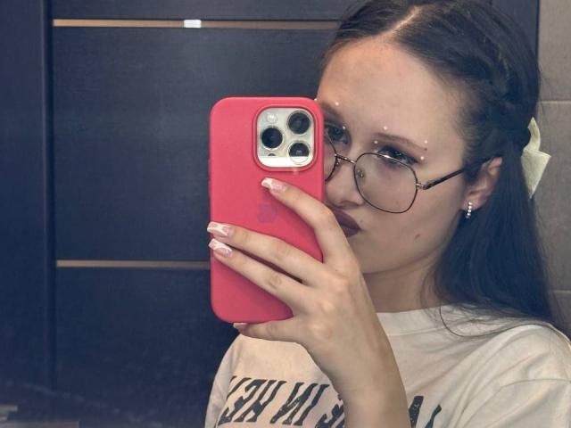 MonaCloudI mit Brille, rosafarbenen Nägeln und einem weißen Hemd, hält ein rosa Handy und macht ein Selfie im Spiegel. Sie hat ein kleines Nasenpiercing und eine weiße Schleife im Haar..