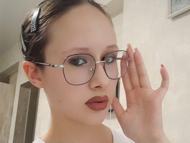 MonaCloud, portant des lunettes, avec une peau claire, des cheveux bruns attachés dans un bandeau, et un vernis à ongles rose, touchant son visage, a un piercing au nez et porte un rouge à lèvres brun, vêtue d'une chemise blanche..
