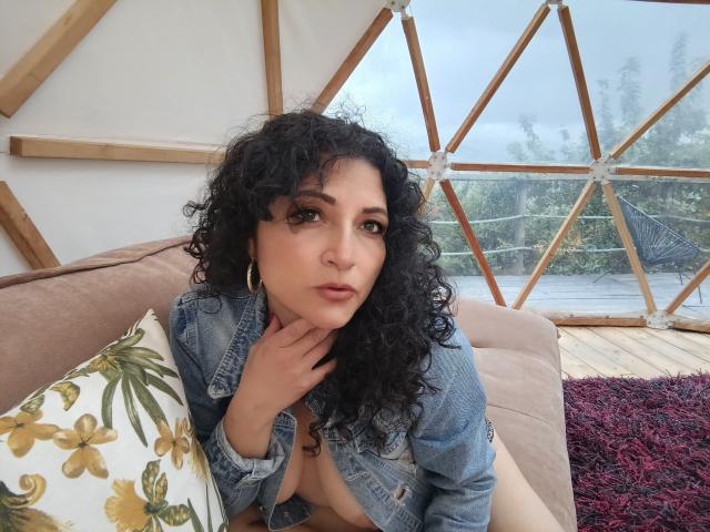 Arianndy with curly black hair, wearing a denim jacket, sits on a beige couch, showing her small tits, hand on neck, gold hoop earrings, floral pillow, and red rug.