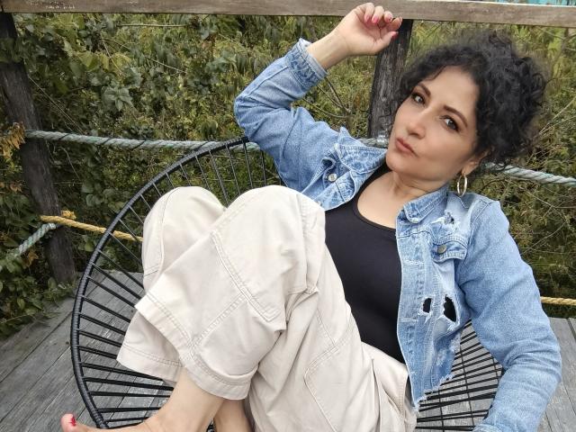 Arianndy with curly black hair, wearing a blue denim jacket, black top, and beige pants, sitting in a black chair, looking up with a relaxed expression. Boobs, ass covered.
