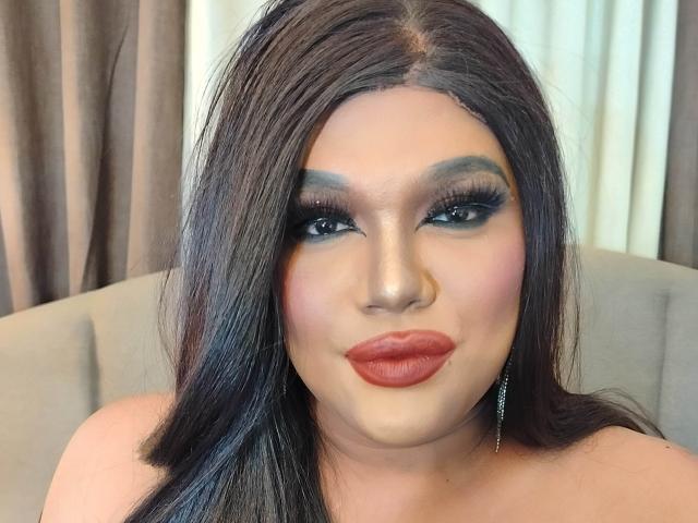 InsatiableSexQueen heeft donker, steil haar, dikke eyeliner en rode lippenstift. Haar borsten zijn licht zichtbaar, en ze draagt een zilveren oorbel. Ze kijkt recht in de camera..