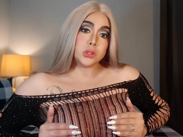UnforgetableMaria with blonde hair, black mesh top, holding her tits, white nails, pink lipstick, and a tattoo on her chest, looking at the camera with pouty lips.