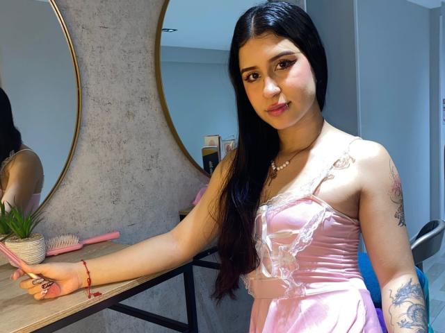 AnnaTorress with long black hair, pink satin dress, and tattoos, holding a pink brush, standing in front of a mirror, looking at camera, wearing a pearl necklace.