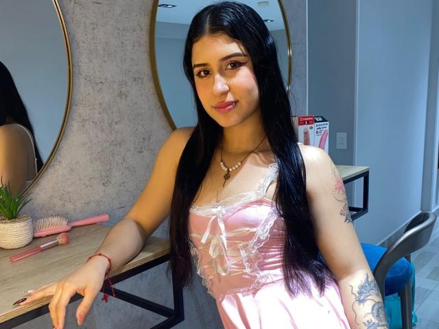 AnnaTorress, con cabello negro largo, vestido de satén rosa, encaje, pequeños senos, collar, tatuaje en el brazo derecho, un pincel rosa, un espejo y un tocador de madera en una habitación muy iluminada..