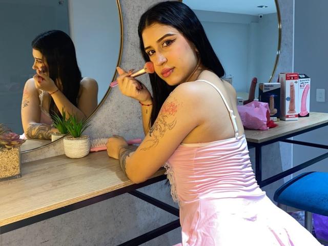 AnnaTorress, con cabello negro, vestido rosa, aplicándose lápiz labial rosa, tatuaje en el brazo, reflejo en el espejo, planta en maceta, bolso de maquillaje y un vibrador sobre una mesa de tocador de madera..