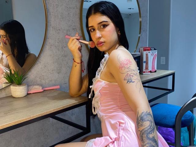 AnnaTorress with long black hair, pink satin dress, and tattoos, applying pink lipstick, showing her pouty lips, sitting on a blue chair, wooden vanity with pink brush.