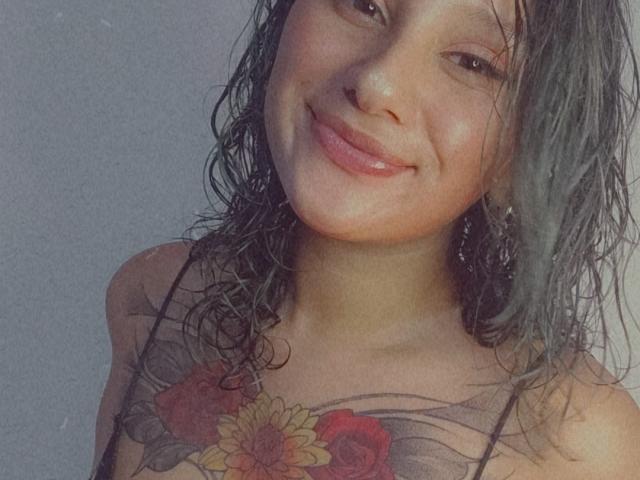 Milanixon with wet, dark green hair, smiling, showing tattoo of sunflower and red roses on chest, wearing thin-strapped top, slightly shiny lips, and dangling earrings.