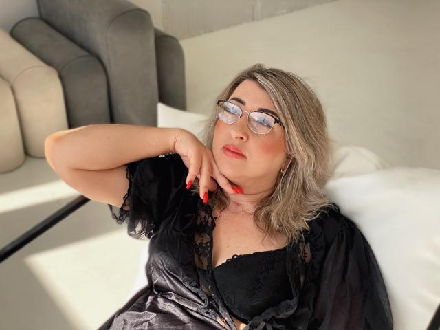 HelenEnigma with glasses, blonde hair, red nails, black lace dress, black bra, lying on bed, hand on neck, looking up, sunlight, grey couch in background.