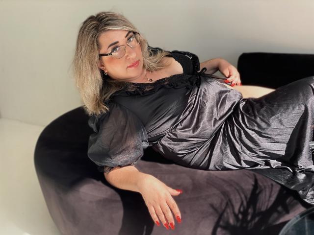 HelenEnigma with blonde hair, glasses, and red nails, laying on a black chair in a shiny black dress, exposing her ass and puffy tits. She's looking to the side.