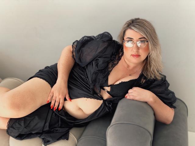 HelenEnigma with glasses, blonde hair, red nails, black lace bra, and open black robe, laying on gray couch, showing her tits and some of her ass.